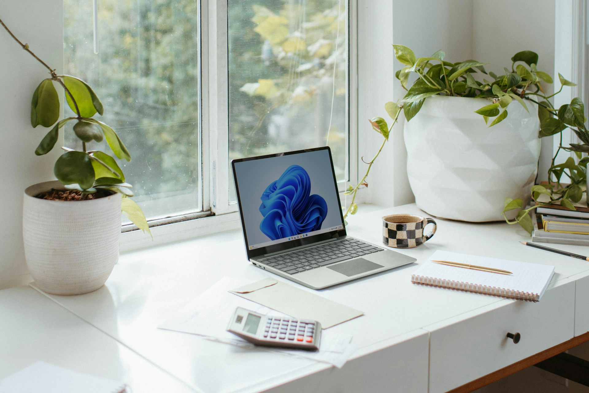 Surface Laptop open on a desk next to a window on a sunny day with papers, a calculator, and a checkered mug on the desk